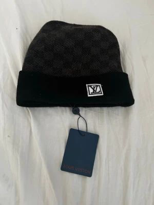 Rutig svart väst från Louis Vuitton - Snygg svart väst från Louis Vuitton med diskret rutigt mönster och bred uppvikt kant. Västen har en vit LV-logga framtill och är tillverkad i mjukt ullmaterial. Perfekt för dig som vill ha en exklusiv och stilren look.