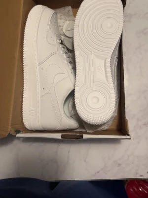 Nike Air Force 1 vita sneakers - Nike Air Force 1 sneakers i helvit färg med klassisk låg siluett. Skorna har snörning, rund tå och platt sula. Tillverkade i skinn med ikonisk Nike-logga på sidan och detaljerade sömmar. Perfekta för dig som gillar stilrena och tidlösa sneakers.