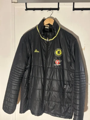 Chelsea pufferjacka från Adidas, svart - Svart pufferjacka från Adidas med Chelsea FC-logga och Carabao-märke på bröstet. Jackan har gul detalj vid kragen, dragkedja framtill och två sidofickor med dragkedja. Perfekt för dig som gillar fotboll och vill ha en sportig stil. lite sliten på adidasloggan.