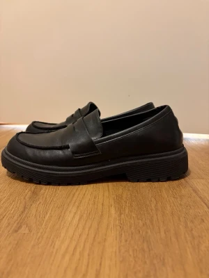 Svarta loafers från Stingers, storlek 40 - Snygga svarta loafers från Stingers i storlek 40. Skorna har en klassisk design med rund tå och platt sula, tillverkade i skinn med en stilren ovandel. Perfekta för dig som gillar en enkel och clean look med chunky sula.