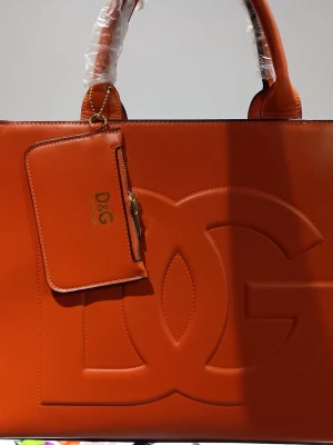 Handväska  - Snygg orange handväska från med stor präglad logga på framsidan och matchande liten plånbok med dragkedja. Väskan har två robusta handtag och är tillverkad i slätt skinnmaterial. Perfekt för dig som gillar stilrena och trendiga accessoarer.