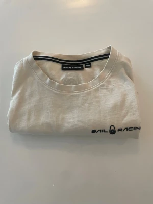 Beige sail racing T-shirt  - Säljer en stilren beige t-shirt från Sail Racing med svart logotyp på bröstet och en mindre logga i nacken. T-shirten har rund hals och korta ärmar, tillverkad i mjuk bomull som känns skön mot huden. Perfekt för dig som gillar enkel och sportig stil. Köpt på kidsbrandstore