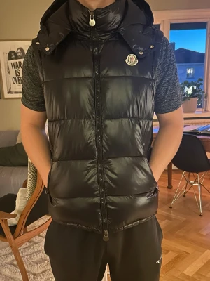 Svart dunväst från Moncler - Säljer en svart, glansig dunväst från Moncler med huva och dragkedja framtill. Västen har klassisk Moncler-logga på bröstet och quiltad design. Perfekt för lager-på-lager och riktigt snygg till streetwear-stil.