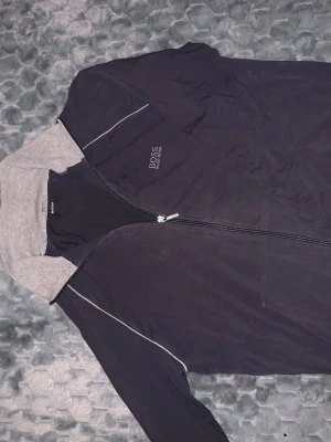 Svart och grå hoodie från Hugo Boss - Säljer en svart hoodie från Hugo Boss med gråa muddar och krage. Tröjan har hel dragkedja framtill, diskret Boss-logga på bröstet och vita detaljer längs ärmarna. Tillverkad i mjuk bomull för skön känsla och stilren look. Sitter mer som S