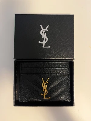 Svart YSL plånbok i skinn - Säljer en stilren svart plånbok från YSL i äkta skinn med guldigt YSL-emblem på framsidan. Plånboken har flera kortfack och ett snyggt quiltat mönster. Perfekt för dig som gillar lyxiga accessoarer och vill ha något som sticker ut.