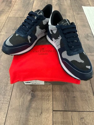 Valentino Garavani Rockrunner sneakers - Snygga Valentino Garavani Rockrunner sneakers i marinblått, svart och vitt med camouflagemönster. Skorna har ovandel i mocka och mesh, samt detaljer i skinn. Klassisk snörning och platt sula för en sportig look. Perfekta för dig som vill sticka ut med exklusiva sneakers.