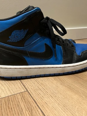 Nike Air Jordan 1 svart/blå sneakers - Säljer ett par Nike Air Jordan 1 sneakers i svart och blått skinn med klassisk hög siluett. Skorna har svarta snören, blå yttersula och ikoniska Air Jordan-loggan på sidan. Perfekta för dig som vill ha en streetwear vibe och gillar retro basketstil.