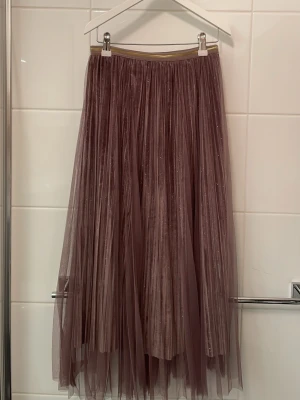 Glittrig långkjol från Winston & Elle - Säljer en superfin långkjol från Winston & Elle i en lila ton. Kjolen är gjord i glittrigt meshmaterial med ett sammet-imiterande innerfoder och har ett elastiskt midjeband med guldrand. Vet ej storlek men pga elastiskt midjeband passar den ungefär alla storlekar.