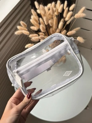 Transparent axelväska Sol de Janeiro - Snygg transparent axelväska från Sol de Janeiro med vit dragkedja och vit justerbar rem med orange text. 