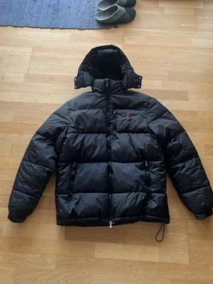 Svart pufferjacka från Polo Ralph Lauren - Svart pufferjacka från Polo Ralph Lauren med röd broderad logga på bröstet. Jackan har huva, två dragkedjeförsedda fickor och justerbar nederkant med snörning. Perfekt för kalla dagar nu i vintern!❄️⛄️
