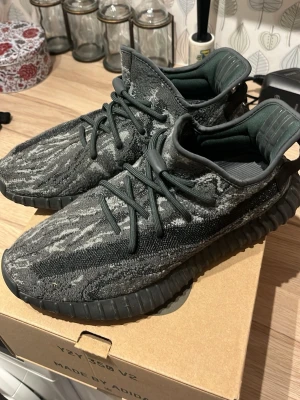 Adidas Yeezy Boost 350 V2 mörkgrå - Säljer ett par Adidas Yeezy Boost 350 V2 i mörkgrå och svart färg med mönstrad ovandel. Skorna har snörning, stickad textil och en platt sula med boost-teknologi. Snygg och unik design som sticker ut med sitt camo-liknande mönster och chunky siluett. Orginalbox medföljer. Skorna har inte heller några tydliga tecken på användning och är sparsamt använda. 