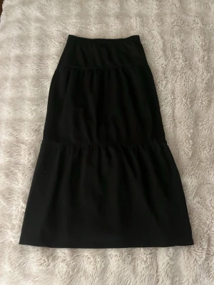 Svart långkjol från Kappahl - Enkel och snygg svart långkjol från Kappahl i storlek 134/140. Kjolen har en rak passform med lätt volang nedtill och bred resår i midjan för extra komfort. Perfekt att styla med en cool topp eller hoodie.