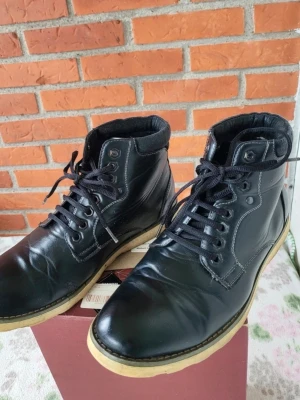 Svarta skinnkängor med snörning 41 passar 42 - Svarta kängor i skinn med snörning och kontrasterande ljus sula. De har rund tå och förstärkt krage i textilmaterial för extra komfort. Snygga sömmar och klassisk design som passar till jeans eller chinos. Perfekt för dig som gillar stilrena och robusta skor.