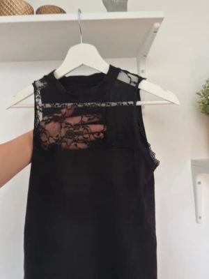 Svart ärmlös topp med spetsdetaljer - Säljer en svart ärmlös topp med transparent spets upptill och runt axlarna. Toppen har en hög halsringning och ett elegant, feminint uttryck tack vare spetsen. Perfekt att styla med både jeans och kjol för en trendig look.