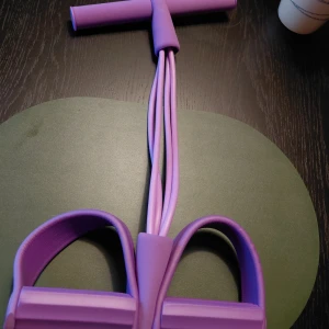 Lila träningsband med fotpedaler - Säljer ett nytt lila träningsband med fotpedaler och handtag, perfekt för hemmaträning. Bandet har fyra elastiska rör och är tillverkat i slitstark syntet. Smidig att använda för helkroppspass och styrkeövningar. Färgen är snyggt lila och designen är modern. Mitt pris: 100:-