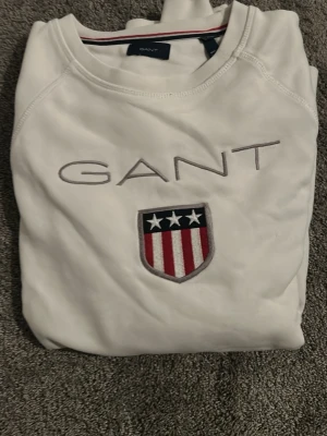 Vit sweatshirt från GANT med emblem - Vit långärmad sweatshirt från GANT med broderad logga och ett stort emblem med stjärnor och ränder på bröstet. Rund hals med snygg röd och marinblå rand på insidan av kragen. Perfekt för dig som gillar klassisk och sportig stil.