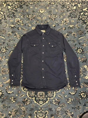 Vintage Overshirt  - Snygg mörkblå overshirt från Sons i storlek L. Skjortan har två bröstfickor med knappar, klassisk krage och tryckknappar längs hela framsidan. Materialet är mjukt och känns som bomull, perfekt att ha över en t-shirt. Passformen är normal och ärmarna är långärmade.