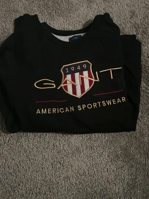 Svart sweatshirt från GANT - Svart sweatshirt från GANT med broderad logga och emblem på bröstet i guld, rött och vitt. Tröjan har rund hals och ribbade muddar vid ärmslut och nederkant. Perfekt för dig som gillar sportig och klassisk stil.