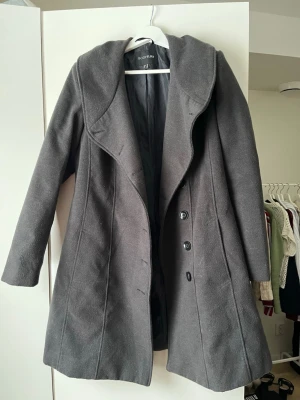 Mörkgrå kappa från Bodyflirt - Snygg mörkgrå kappa från Bodyflirt med bred krage och knappar framtill. Jackan har en feminin siluett och är perfekt för kyliga dagar. Materialet är mjukt och den har långa ärmar för extra värme. Hängöglan på jackan har gått upp men går lätt att sy fast igen.