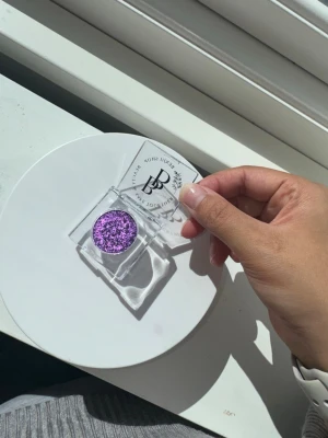 Lila glitterögonskugga från Benjii shop - Superfin lila ögonskugga med intensivt glitter i en rund dosa med transparent lock. Perfekt för att skapa en riktigt glittrig och färgstark look. Skuggan har en krämig textur och kommer i en snygg förpackning med logga på locket.