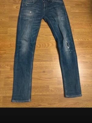 Blå jeans från Dondup med slitning - Säljer ett par blå jeans från Dondup med snygga slitningar och tvättade detaljer. Klassisk femficksmodell med raka ben och kontrastsömmar. Jeansen har en brun patch med Dondup-logga bak i midjan och en unik broderad detalj på bakfickan. Tveka inte på att skriva om ni undrar något.