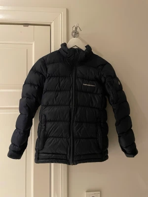 Svart Peak Performance pufferjacka 160 - Svart pufferjacka från Peak Performance med hög krage och dragkedja framtill. Jackan har ett quiltat mönster och två sidofickor. Perfekt för kalla dagar och har en clean, sportig look som funkar till det mesta. Ny pris 4100kr mit 999kr