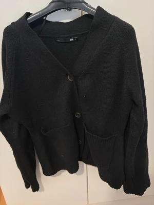 Svart stickad kofta från 157 - Svart stickad kofta från 157 i storlek S. Koftan har v-ringad hals, tre knappar framtill och två rymliga fickor. Perfekt att slänga på sig när det är lite kyligt ute. Enkel och stilren design som funkar till många olika outfits.