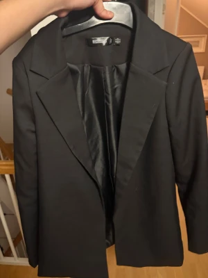 Svart kavaj från Missguided, strl 34 - Stilren svart kavaj från Missguided i storlek 34. Klassisk modell med breda slag, helfodrad insida och lång ärm. Perfekt för dig som vill ha en clean och snygg look. Materialet är slätt och känns bekvämt mot huden.