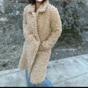 Beige teddyjacka från exklusiva Soulmate stl.XS - Säljer en lång, fluffig teddyjacka från Soulmate i beige färg. Jackan har stora lockar, klassisk krage och knäppning framtill. Perfekt för dig som vill ha en mysig och trendig look under kyliga dagar.