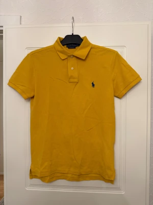 Gul pikétröja från Ralph Lauren - Säljer en snygg gul pikétröja från Ralph Lauren med klassisk krage och två knappar framtill. Tröjan har korta ärmar och den ikoniska blå Ralph Lauren-loggan broderad på bröstet. Perfekt för dig som gillar stilrena och färgglada plagg.