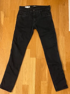 Svarta Replay Anbass jeans slim fit - Svarta Replay Anbass jeans med slim fit och klassisk femficksdesign. Jeansen har svarta sömmar, Replay-logga på fickan och läderpatch bak i midjan. Tillverkade i stretchig bomullsdenim för en bekväm passform.