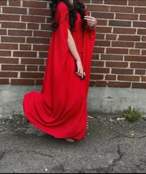 Röd långklänning med capeärm - Säljer en röd långklänning med capeärmar och elegant, svepande passform. Klänningen har en lös siluett och är perfekt för dig som vill sticka ut med en dramatisk look. Passar dig som gillar färgstarka och unika plagg.