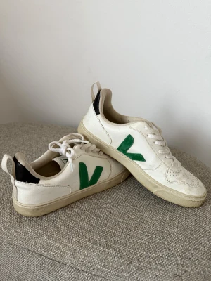 Veja vita sneakers med grönt V - Säljer ett par klassiska Veja sneakers i vitt skinn med grönt V på sidan och svart detalj vid hälen. Modellen har rund tå, perforerat mönster på tån och platt sula. Skorna har snörning och är låga, vilket ger en clean och fräsch look.