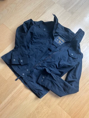 Woolrich fieldjacket  - Snygg svart vindjacka från Woolrich med hög krage, dragkedja och tryckknappar. Jackan har fyra fickor framtill, figurnära passform och är tillverkad i polyester. Perfekt för dig som gillar stilren och sportig look. Sitter som xs! 