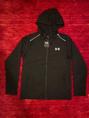 Svart vindjacka Under Armour - Under Armour jacka i hög kvalitet. Lätt och bekväm modell med huva, perfekt för träning eller vardag. Jackan är i superskick och nästan som ny. Det finns en liten lagning i tyget mitt på jackan, men när dragkedjan är stängd syns den knappt alls. I övrigt är jackan i mycket fint skick och håller den höga kvalitet man kan förvänta sig av Under Armour. 🌴🔋🌟 Stolek S/M