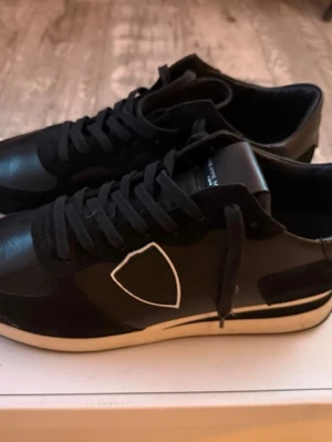 Svarta sneakers från Philippe Model - Snygga svarta sneakers från Philippe model med vit sula och vit detalj på sidan. Skorna har snörning och är tillverkade i skinn och syntet. Perfekta för dig som gillar stilrena och sportiga skor med en modern touch.