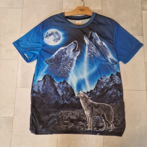 Blå t-shirt med vargar och måne - Säljer en blå t-shirt från The Mountain med ett stort tryck av ylande vargar under fullmåne och berg i bakgrunden. T-shirten har rund hals och korta ärmar. Materialet är mjuk polyester som känns skönt mot huden. Perfekt för dig som gillar djurmotiv och coola prints.