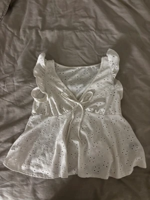 Vit blus med volang och knyt - Supersöt vit blus med broderade hålmönster, volangdetaljer på axlarna och v-ringning med knyt framtill. Blusen är ärmlös och har en lätt utsvängd passform som ger en luftig känsla. Perfekt för varma dagar och en fräsch look.