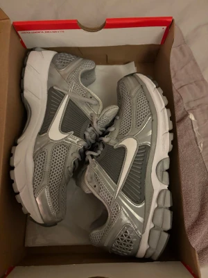 Nike Zoom Vomero 5 Wolf Grey - Nike Zoom Vomero 5 sneakers i färgen Wolf Grey/White. Skorna har en chunky sula, mesh-paneler för extra ventilation och detaljer i syntetmaterial. Swoosh-logga i vitt på sidan och snörning framtill. Perfekt för dig som gillar sportig och modern stil.