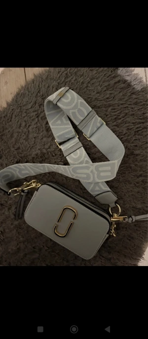 Ljusgrå axelväska Marc Jacobs - Snygg ljusgrå axelväska från Marc Jacobs med bred justerbar axelrem med logga. Väskan har guldfärgade metalldetaljer, dubbla dragkedjor och en dekorativ tofs. Tillverkad i skinn med stilren, boxig form och diskret fack på baksidan.