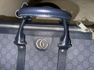 Gucci blå resväska med GG-mönster - Snygg blå resväska från Gucci med klassiskt GG-mönster och silverfärgade detaljer. Väskan har både handtag och justerbar axelrem, dragkedja upptill samt medföljande lås och nycklar. Tillverkad i canvas och skinn för en lyxig känsla.