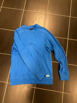 Blå sweatshirt från 157, storlek M - Snygg blå sweatshirt från 157 i storlek M. Tröjan har rund hals, långa ärmar och ribbade muddar vid ärmslut och nederkant. Perfekt för dig som gillar enkel och stilren design. Materialet är mjukt och skönt, troligtvis bomull eller polyester. Använt fåtal gånger!