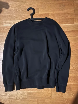 Svart sweatshirt från Weekday, M - En stilren svart sweatshirt från Weekday i storlek M. Tröjan har rund hals, ribbade muddar och är tillverkad i mjuk bomullsmix. Perfekt för dig som gillar enkel och clean look. Passar dig som vill ha en klassisk och bekväm tröja.