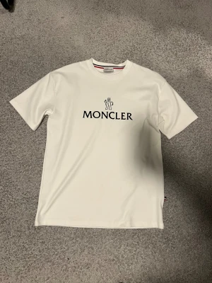 Vit Moncler t-shirt med logga - Vit t-shirt från Moncler med klassisk svart logotyp tryckt på bröstet. Rund halsringning med snyggt röd och marinblå band på insidan av kragen. T-shirten har korta ärmar och är gjord i mjuk bomull för en skön känsla. Perfekt för dig som gillar stilrena designer.