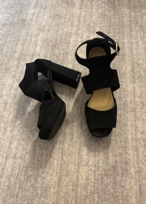 Svarta klackar - Säljer ett par svarta klackar med bred klack och plattform. Skorna har öppen tå och ett snyggt spänne runt ankeln. Materialet är mocka som ger en lyxig känsla och passar perfekt till dig som gillar statement-skor.