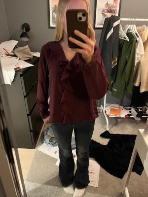 Vinröd blus med volangdetalj - Vinröd blus med långa ärmar och snygga volangdetaljer längs ringningen och framtill. Blusen har en lös passform och är tillverkad i ett lätt, flowy material som ger en skön känsla. Perfekt för dig som vill ha en chic och trendig look.