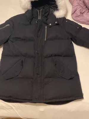 Svart Moose Knuckles dunjacka med päls - Svart dunjacka från Moose Knuckles med vit päls på huvan, flera dragkedjor och snygga detaljer på ärmen. Jackan har långa ärmar, praktiska fickor och broderad insida med hockeymotiv. Perfekt för kalla dagar och har en cool streetstil.