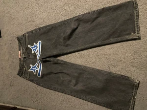 Protect jeans med blå tryck, storlek S - Svarta jeans från Protect i storlek S med coolt blått och vitt tryck på både framsidan och bakfickan. Jeansen har raka ben, kontrastsömmar i gult och är tillverkade i 100% bomull. Perfekt för dig som vill sticka ut med en unik streetwear-look.