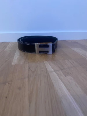 Svart skärp med H-spänne i metall - Snyggt svart skärp med ett stort H-format spänne i silverfärgad metall. Skärpet har vita sömmar längs kanterna och är gjort i slätt skinn med en lätt textur. Perfekt för att ge outfiten en clean och stilren touch.