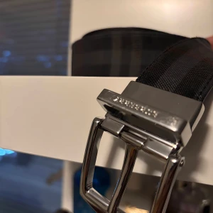  Burberry bälte - Budgivning - Säljer ett snyggt Burberry bälte i svart med diskret rutigt mönster och silverfärgat spänne där Burberry-loggan är ingraverad. Bältet är tillverkat i slitstark syntet med detaljer i skinn och levereras med originalpåse, box och kvitto.       Lägg bud 